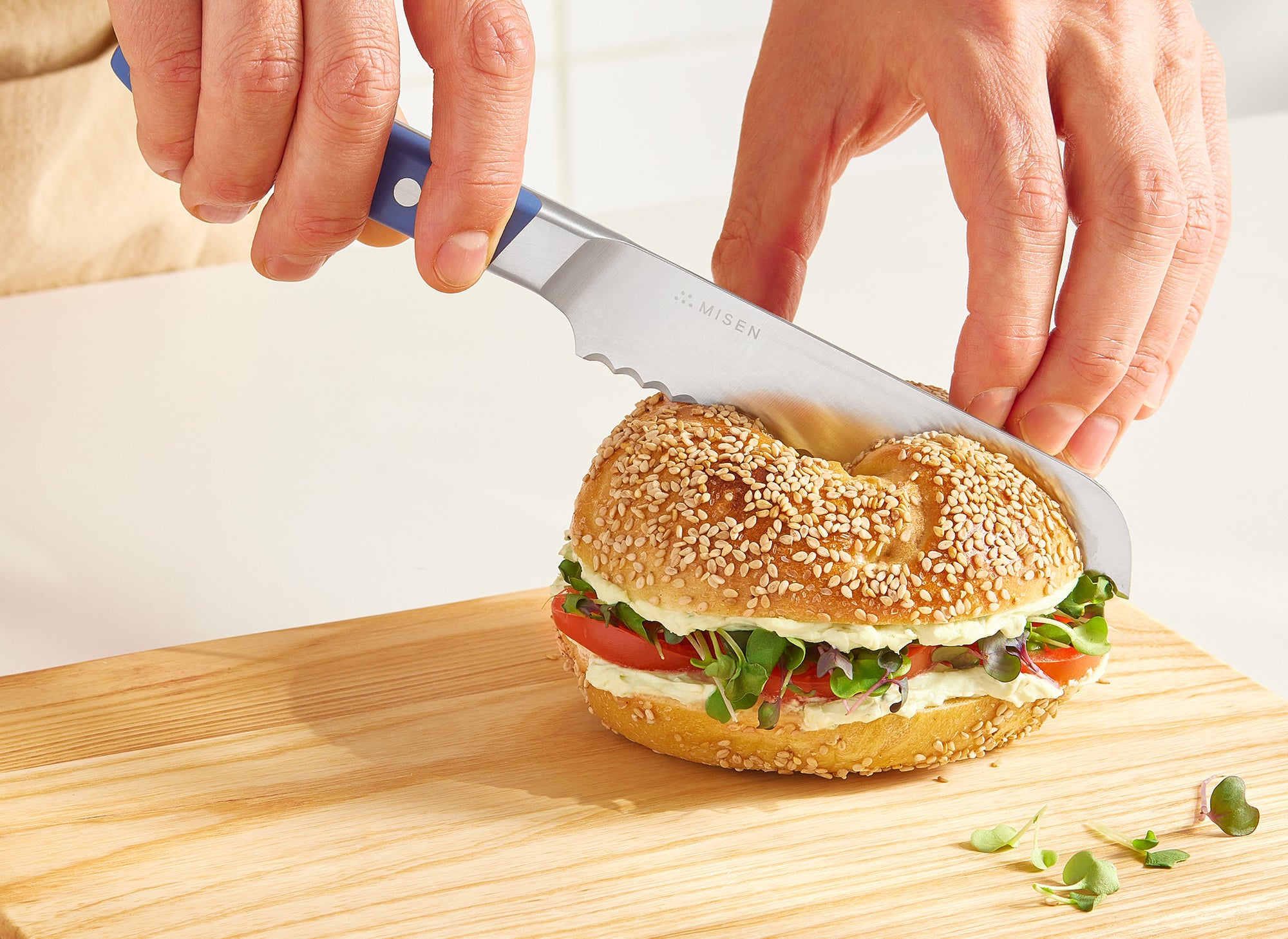 A hand uses a blue Misen Short Serrated Knife to slice a sesame bagel sandwich in half on a Misen Wood Cutting Board. 