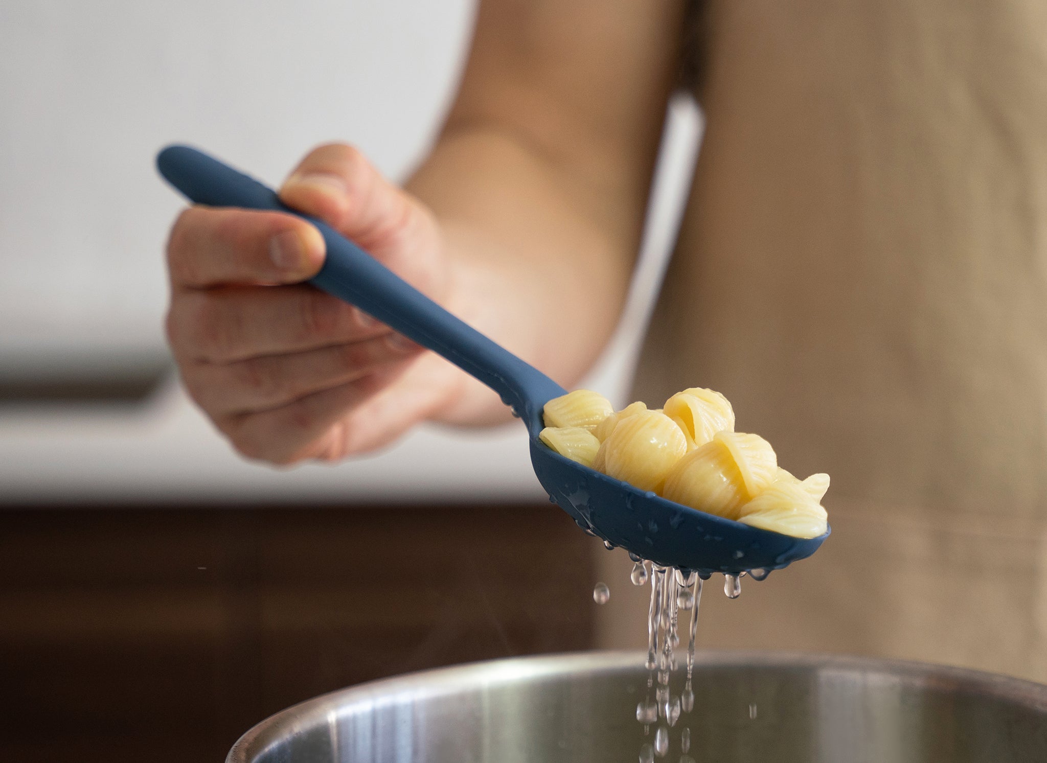 {{blue,black,gray}} A hand lifting cooked shell pasta from a steel pot with a Blue Misen Slotted Spoon, with hot water draining through the spoon.