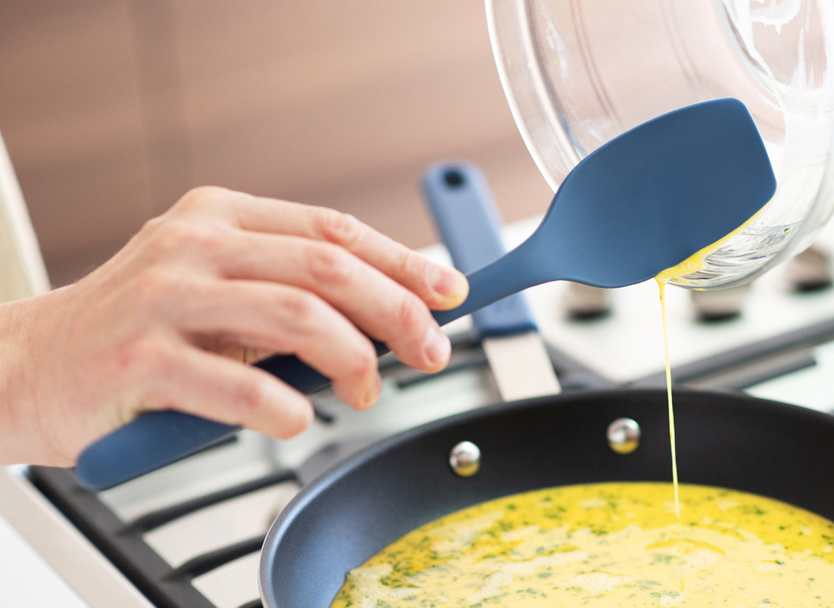 {{blue,black,gray}} A Blue Misen Spoontula scooping liquid scrambled egg from a clear mixing bowl into a nonstick pan on a stovetop.