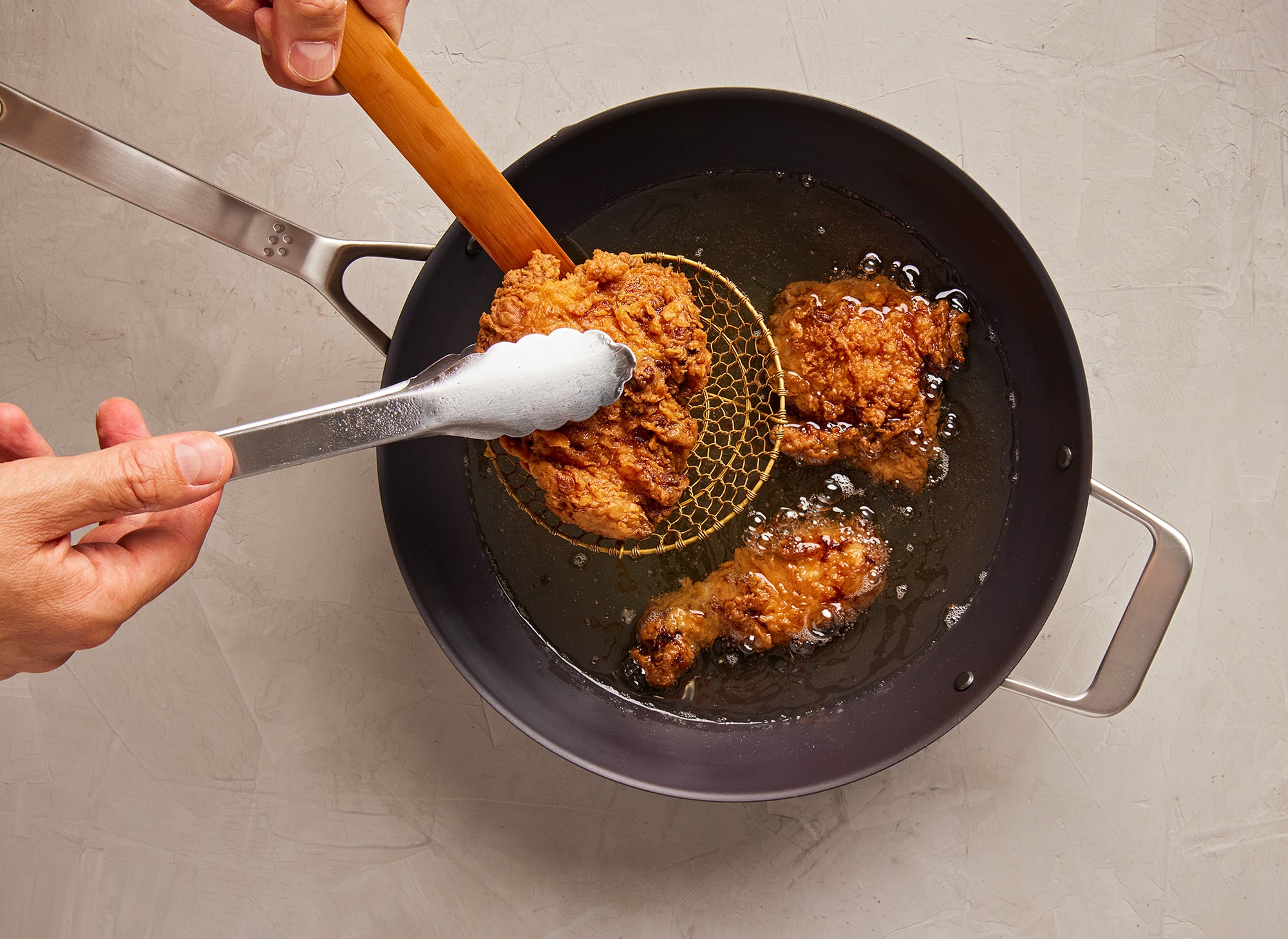 {{12-inch}} A bird’s eye view of a chef pulling a piece of fried chicken out of the Misen Carbon Steel Wok using tongs and a strainer. Additional pieces of chicken can be seen frying in oil in the wok.