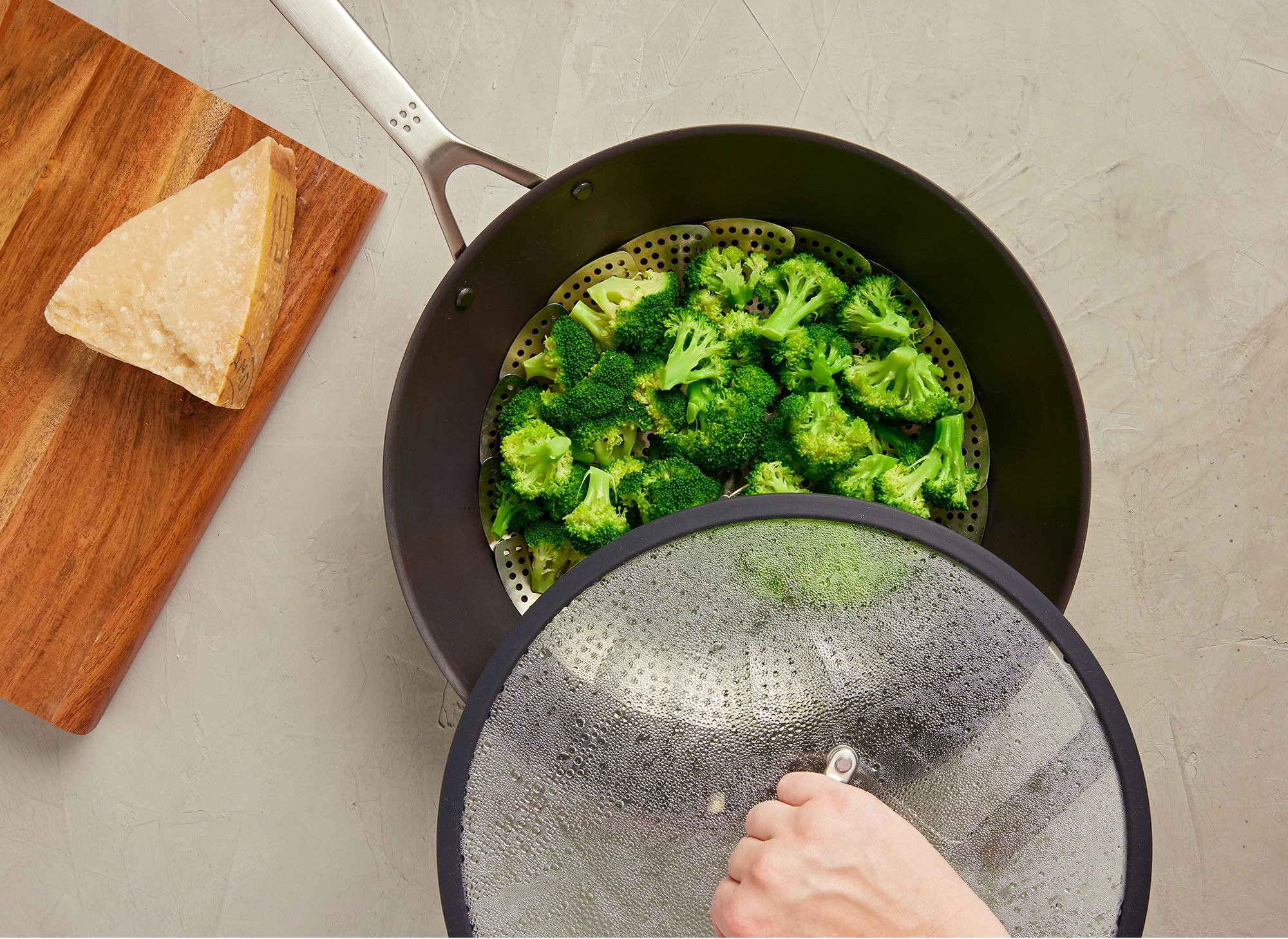 {{12-inch}} A bird’s eye view of the Misen Carbon Steel Wok being used to steam broccoli, which sits in the wok in a metal colander. A chef’s hand removes a steamy lid from the wok. A cutting board with a wedge of cheese can be seen next to the wok.